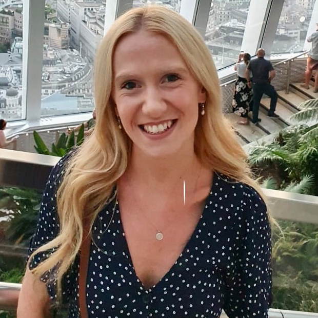 headshot of  Emily Cooke. She is smiling in an office near a staircase, plants and window.