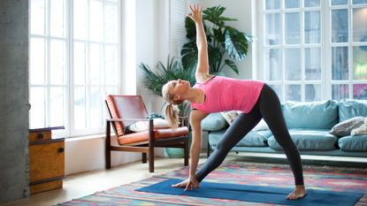 A woman performs a yoga triangle pose at home on a mat. She is in a wide legged stance with her legs straight, her left foot pointing forward and her right foot pointing out. Her torso is twisted towards the camera and she rests one hand on the floor while the other points upwards. Behind her we see a couch, potted palm and leather seat.