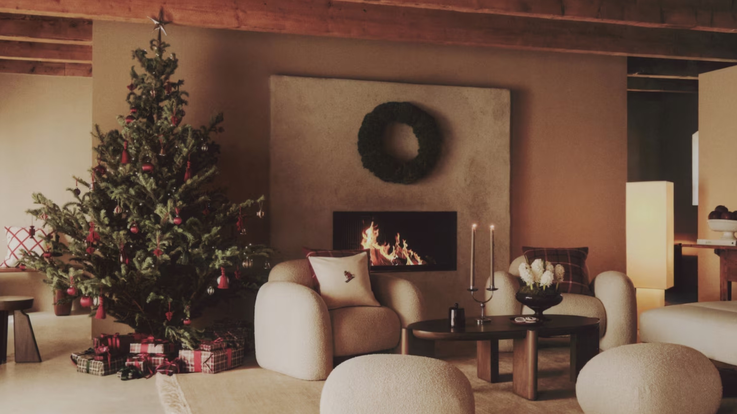 A living room with exposed beams and a lit fireplace with a decorated Christmas tree and a wreath hung above the fireplace