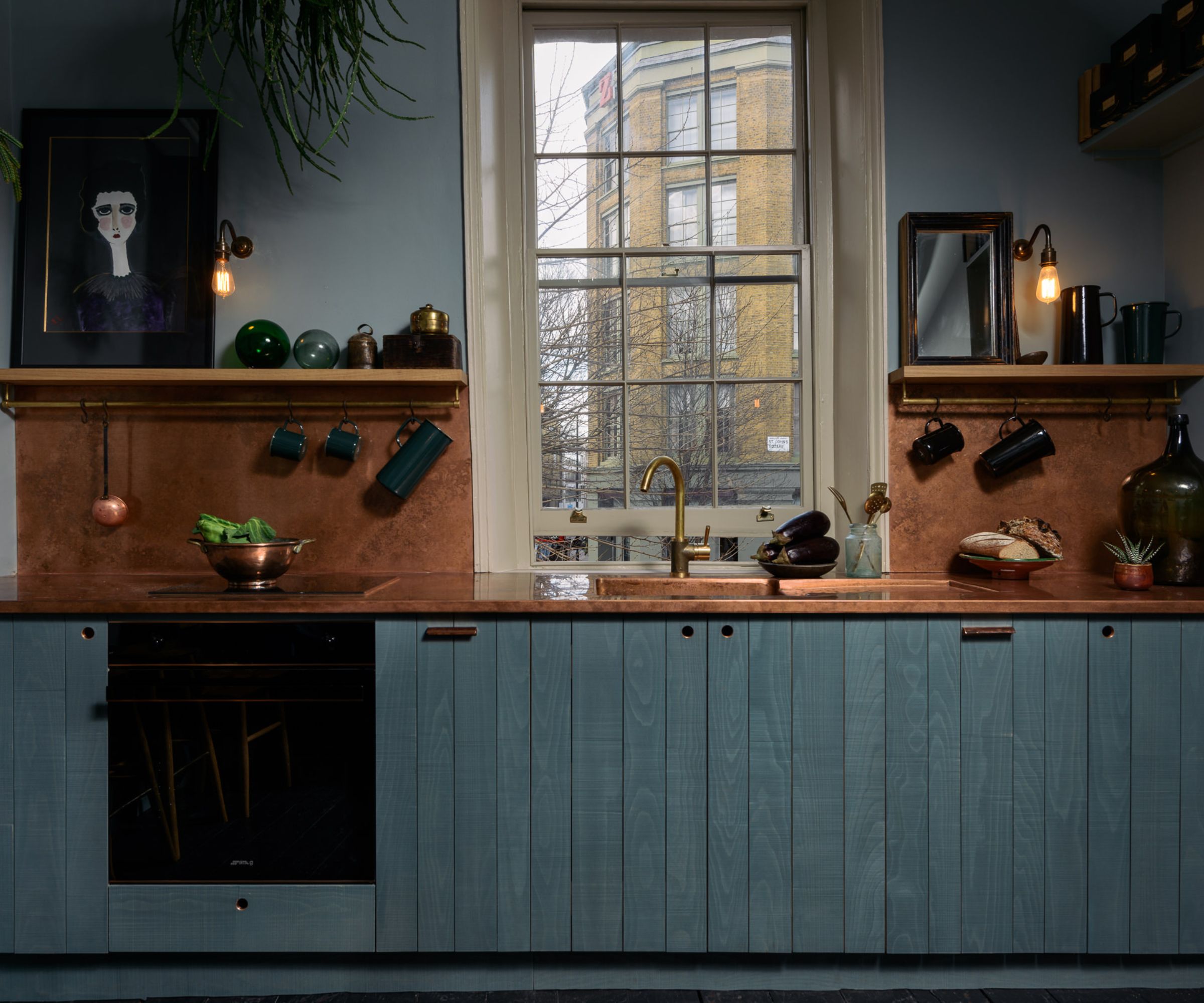 A one-wall kitchen with blue cabinets, copper countertops, and open shelving filled with art and kitchenware