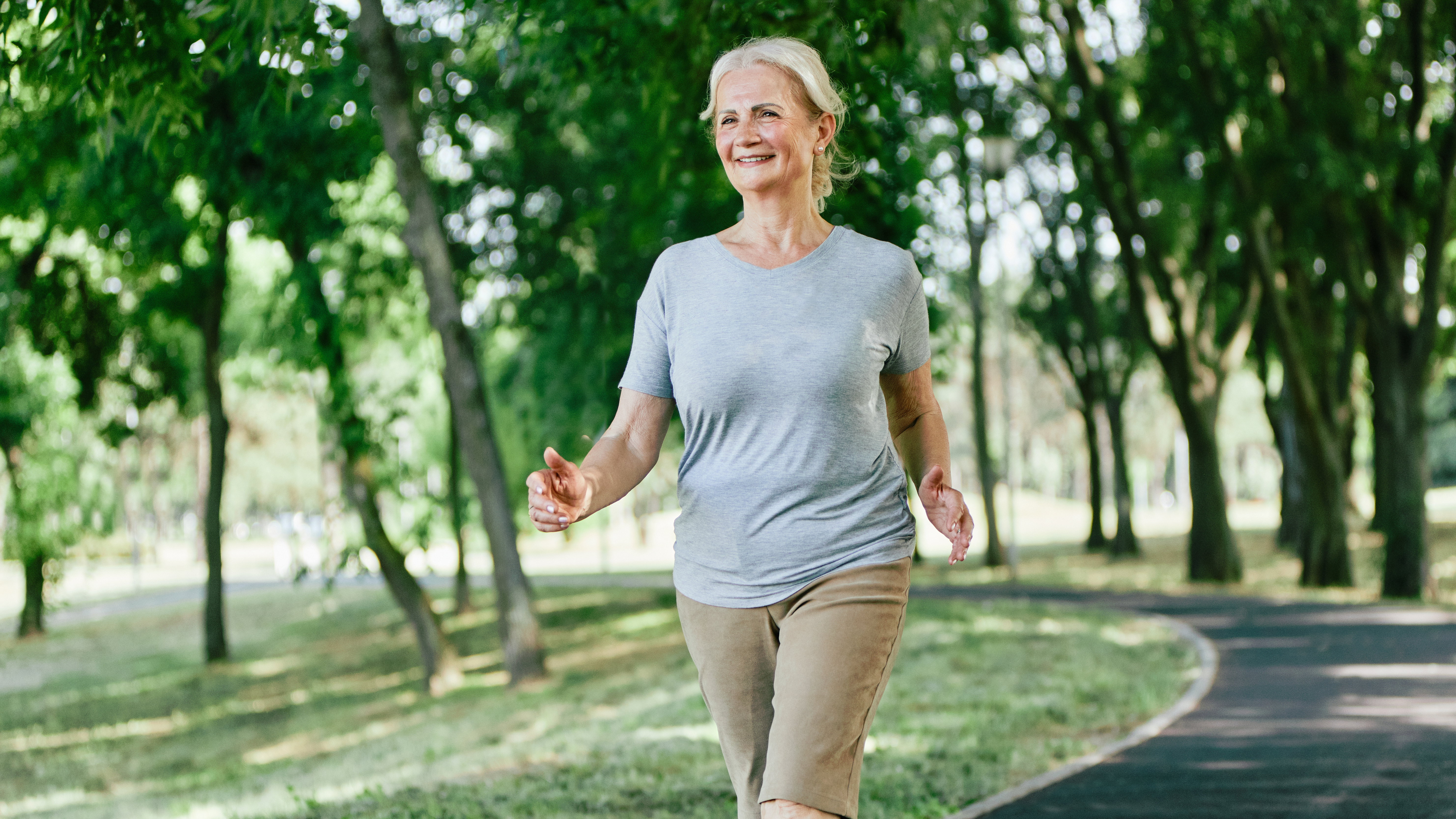 A woman in a t-shirt and trousers walks through a park, her arms swinging by her sides.