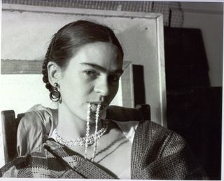 a woman (frida kahlo) holds a necklace in her mouth while sitting in front of a window