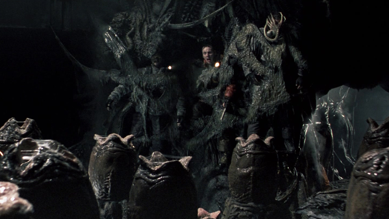 Screenshot from the movie Alien vs. Predator. Three terrified humans have been pasted to a cave wall and are stuck there. Before there is a room full of opening xenomorph eggs. One poor human has a facehugger alien stuck on their face.