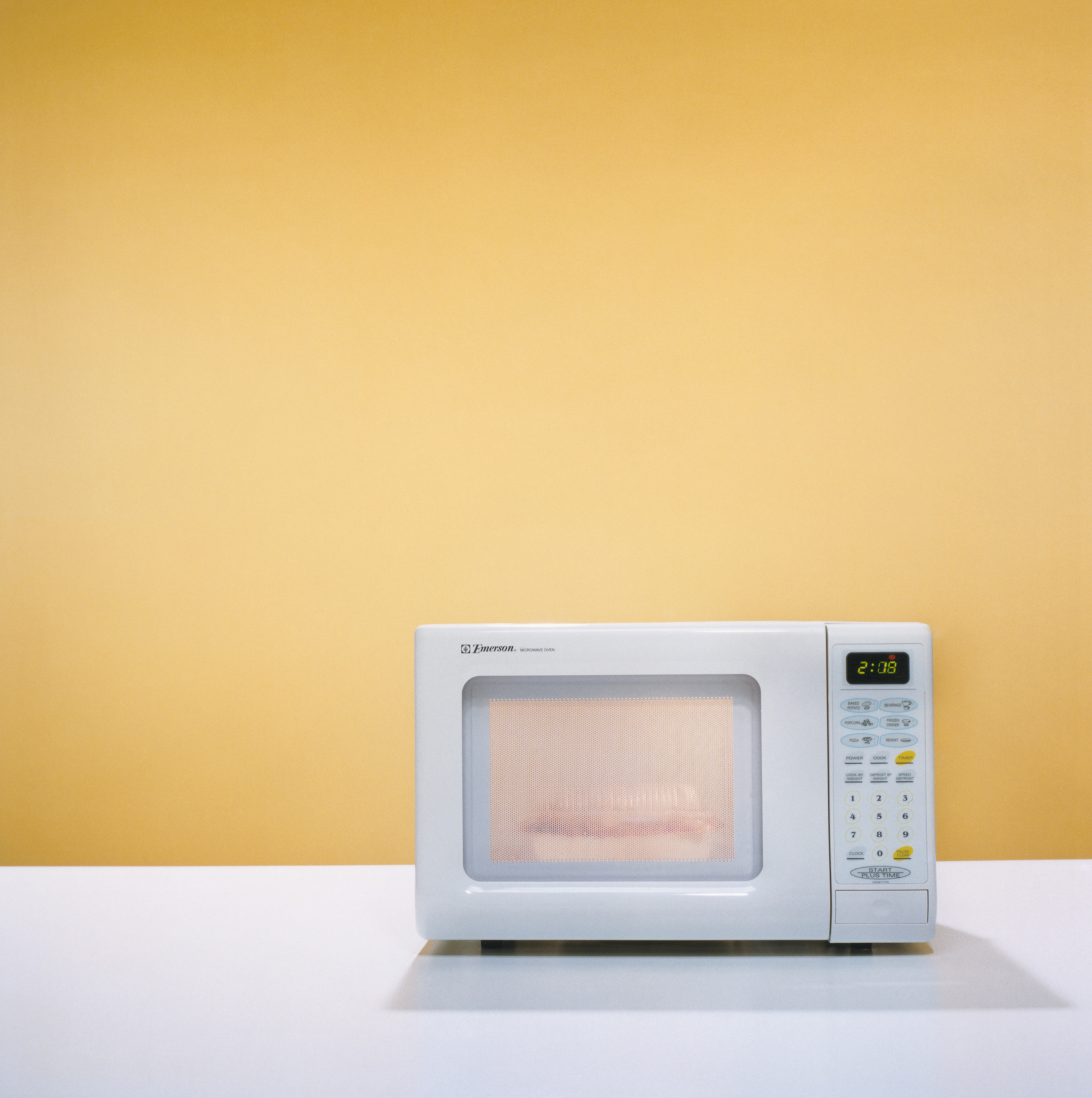 A white microwave sat on top of a white surface against a yellow wall.