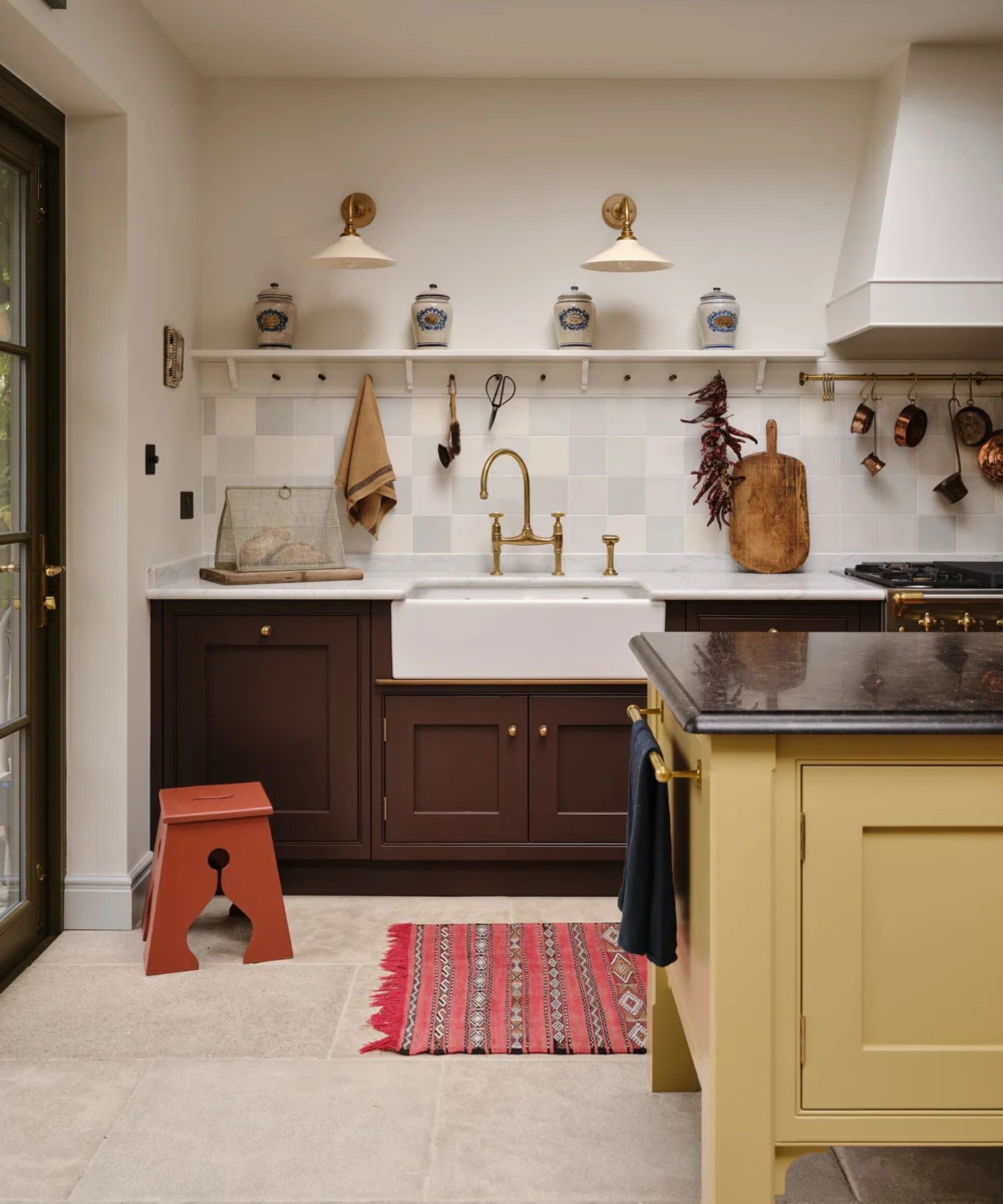 Country kitchen with dark brown lower cabinets, a white farmhouse sink, a yellow island, a red patterned rug, and a shelf displaying ceramic jars under twin sconces