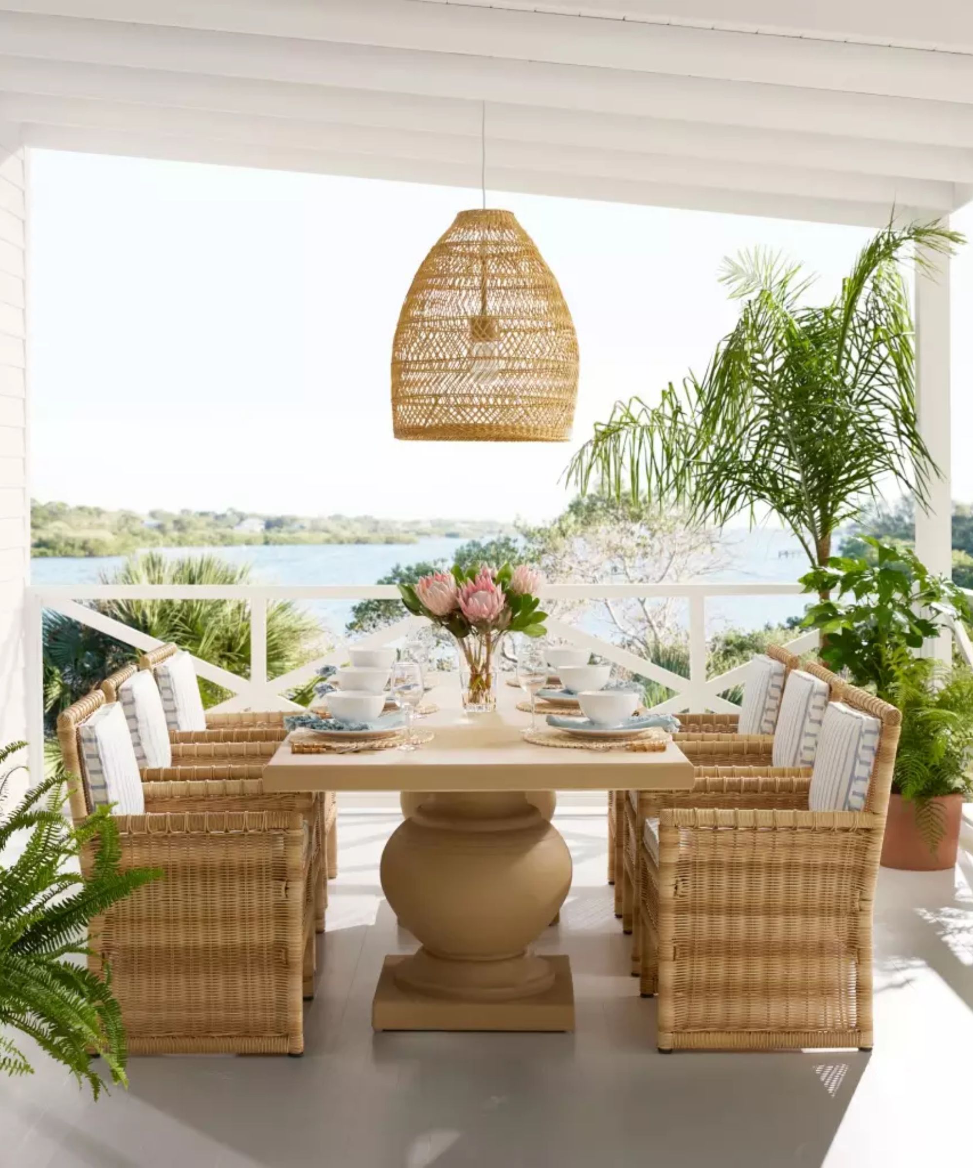 coastal garden veranda with a sea view by serena and lily with a large pedestal wooden table with bamboo and wicker armchairs and a seagrass ceiling pendant