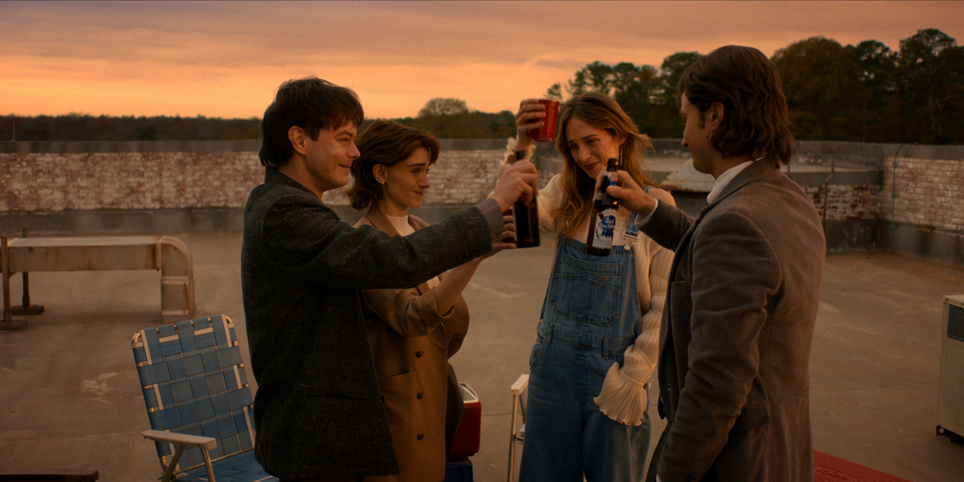 Charlie Heaton as Jonathan Byers, Natalia Dyer as Nancy Wheeler, Maya Hawke as Robin Buckley, and Joe Keery as Steve Harrington in Stranger Things: Season 5 finale cheersing drinks on a rooftop