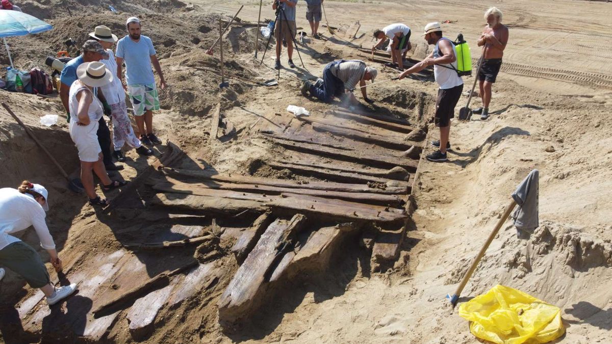 We see a group of people standing around the ancient wooden planks of a boat in a desert-like place.