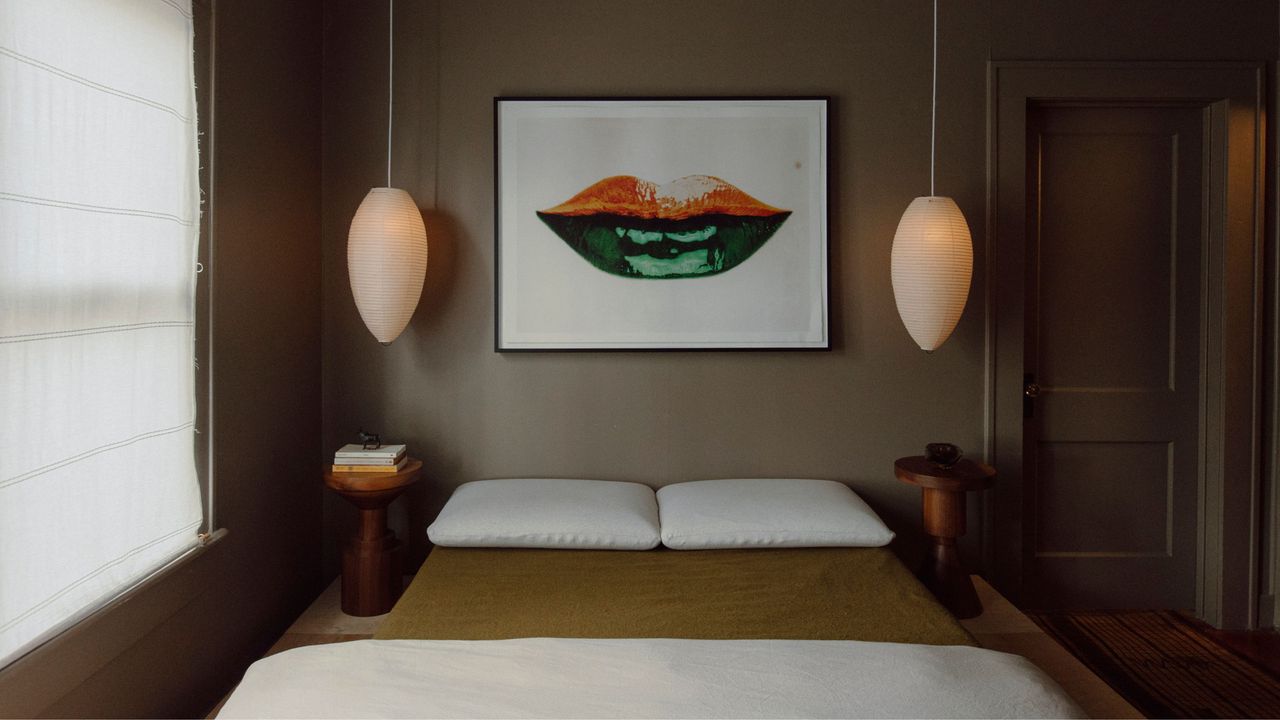 bedroom with tan walls, two pendant lights, roman blinds, and artwork above bed