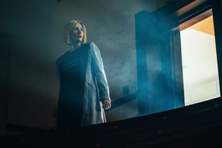The Doctor (Jodie Whittaker) stands in a darkened room with smoke swirling around her and an open door behind her with a light shining through it. The Doctor is looking over her shoulder apprehensively