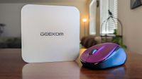 A sleek, white Geekom device sits on a wooden table next to a purple and blue computer mouse. The background features a cozy, softly-lit room with blinds.