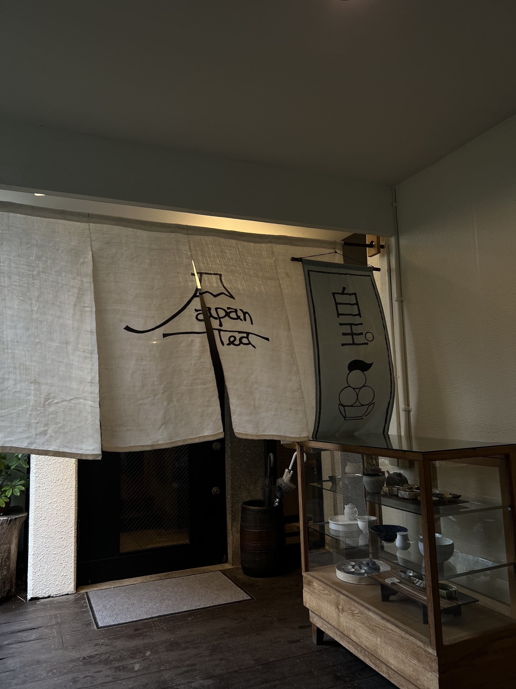 An indoor/outdoor room with white walls, wooden flooring and a glass cabinet full of trinkets. There is a doorway with four white noren curtains in front of it.