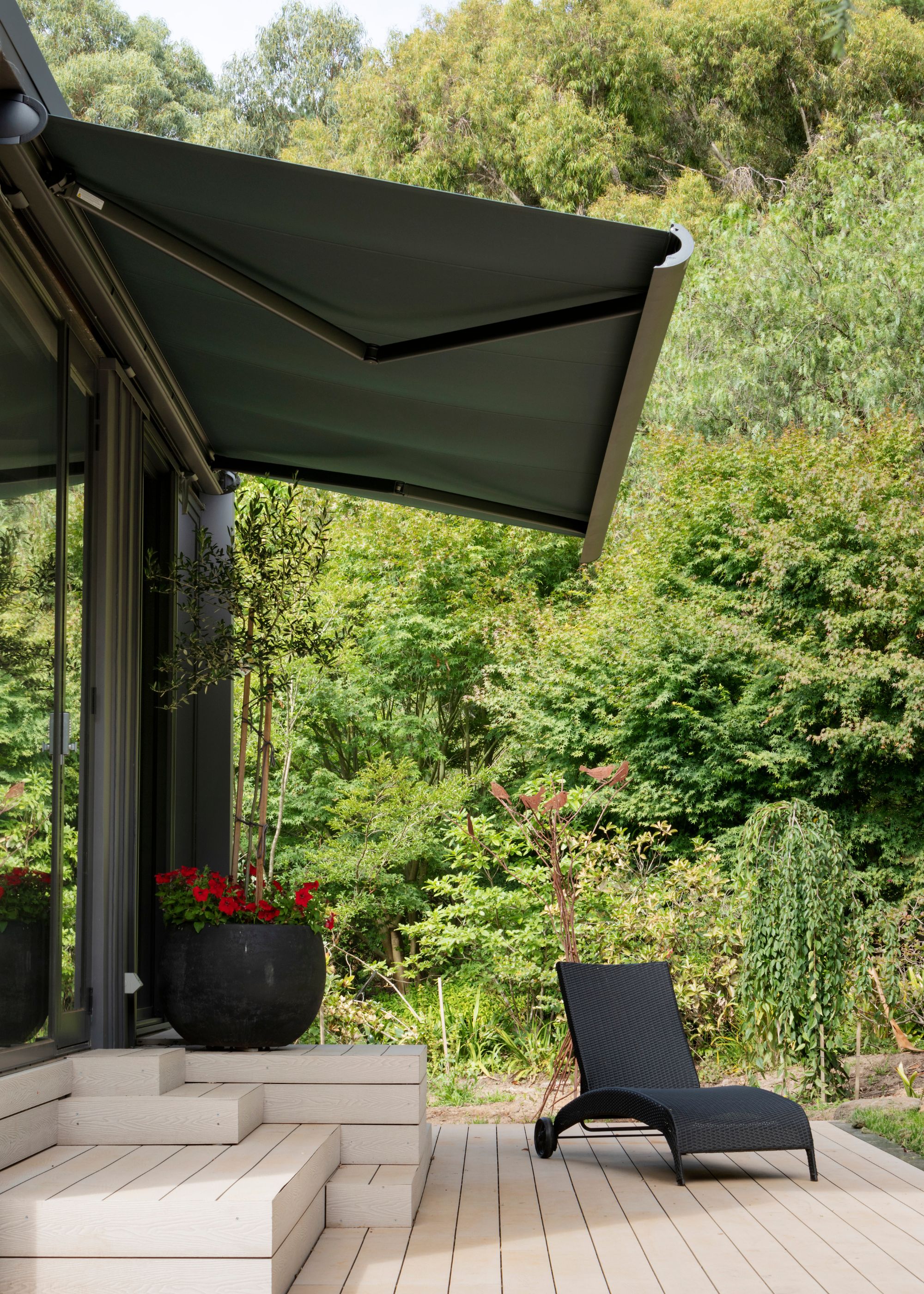 An awning above a patio chair and planter with red flowers in it