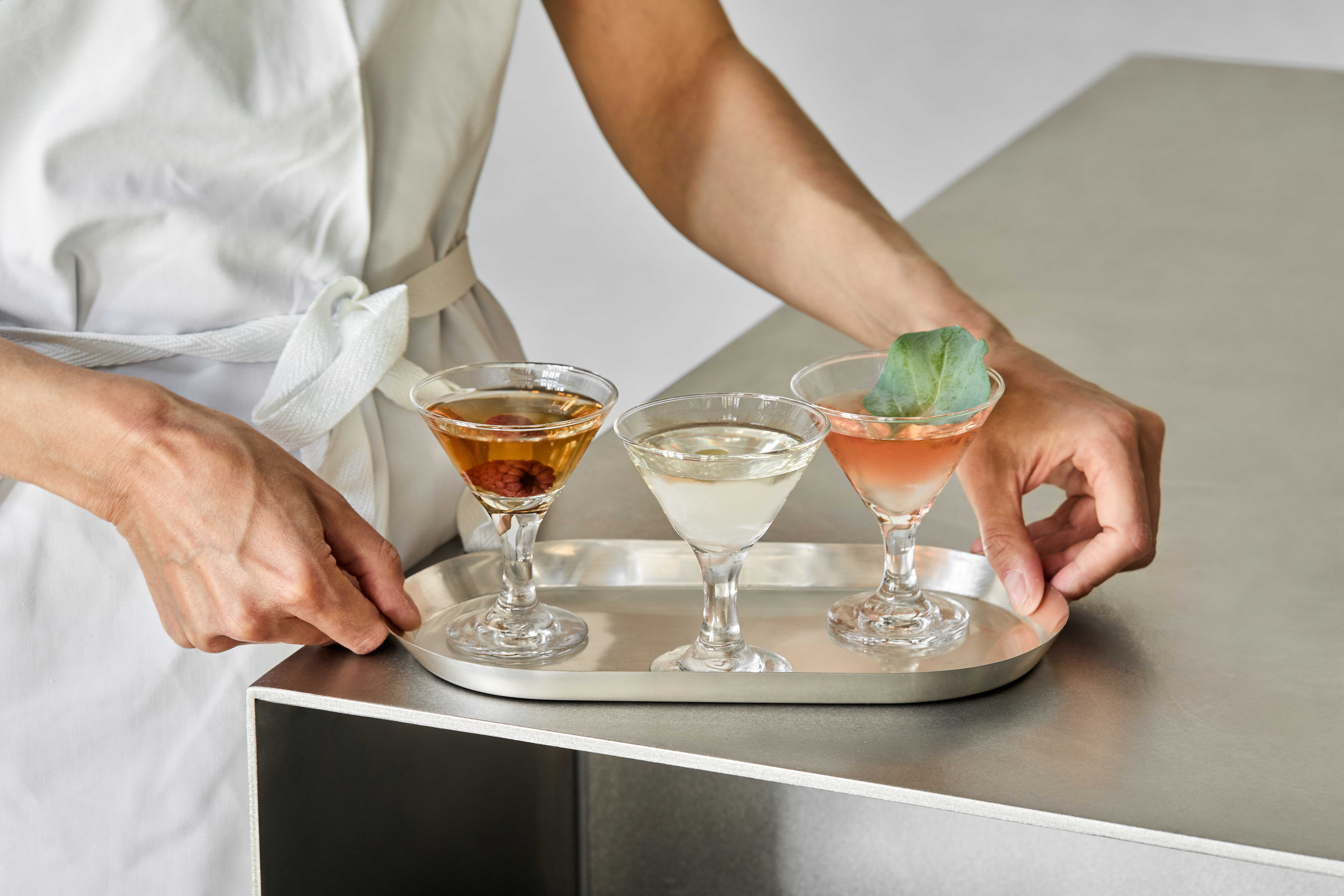 A series of beautifully presented, colorful restaurant plates and cocktails come served in white or chrome tableware and are placed atop silver tables.
