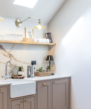 corner of kitchen with butler sink, mushroom coloured base units, small skylight in sloped ceiling, long armed brushed nickel wall light and small black table lamp on corner of worktop