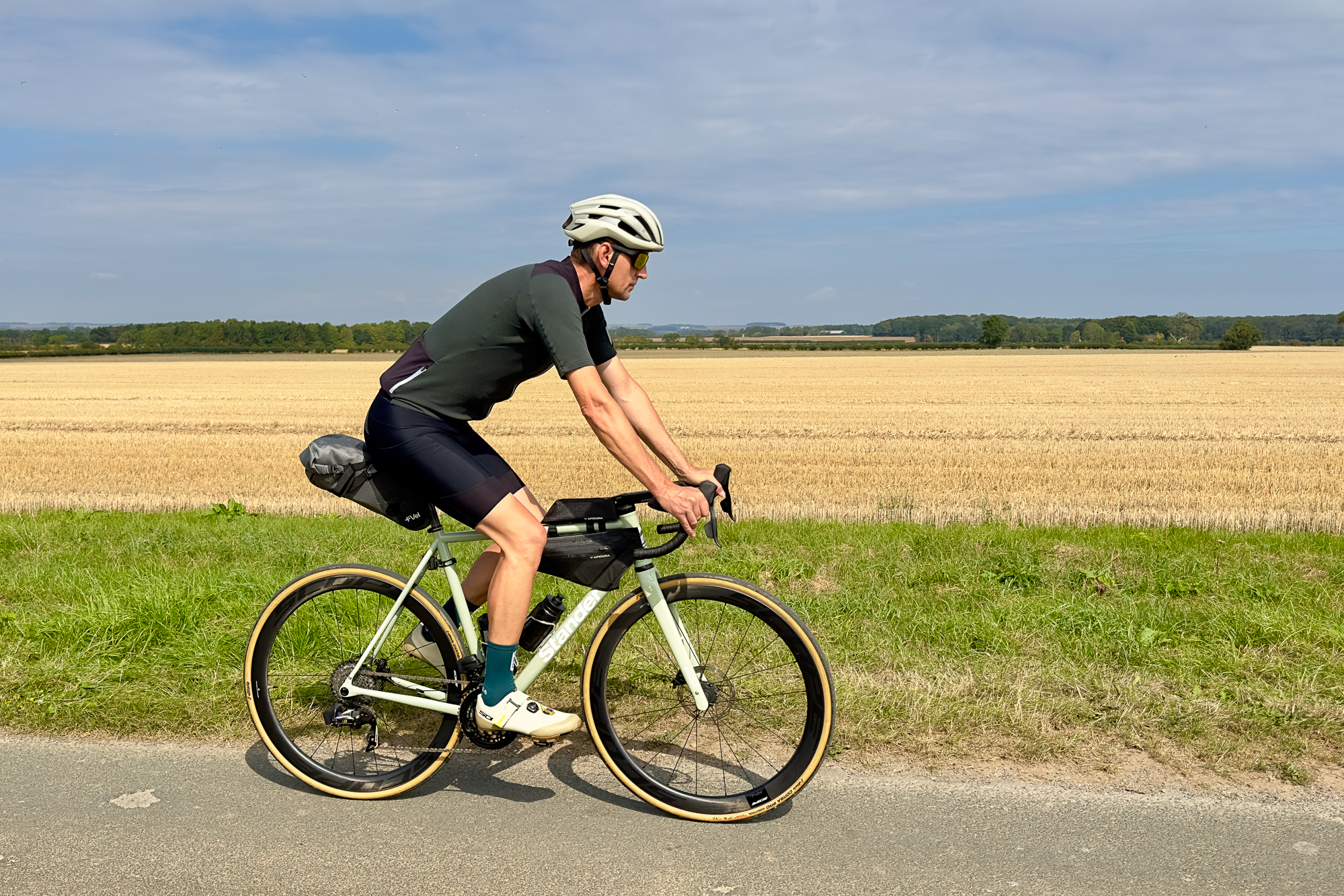 man wearing a dark green jersey and beige helmet on a pale green road bike riding past an arable field