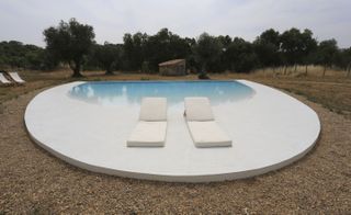 A white oval cement area with a built-in pool and two white loungers in front of it