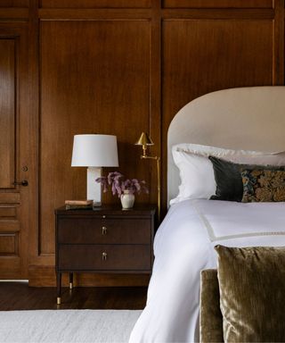 vintage bedroom with wooden panelled walls decorated with a neutral large headboard, white linens and a tapestry covered pillow