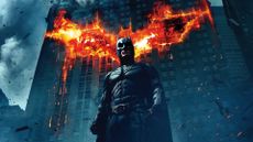 A promo image for The Dark Knight, aka one of the best superhero movies, which shows Batman standing in front of a building with a burning bat logo on it