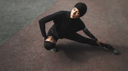 Sweaty Betty to introduce activewear hijab. Young muslim fitness woman in hijab doing stretching exercises outdoors. Dark background with empty place and copy space. Sports concept. Warm up before running training. Black sports clothes. Traditional eastern modest clothing. Healthy lifestyle. Sunny weather.