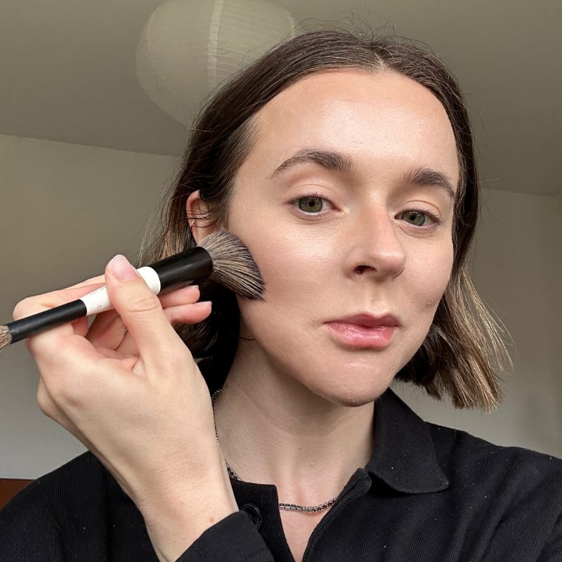 A selfie of Grace Day using a makeup brush to apply a Trinny London bronzer to her cheeks