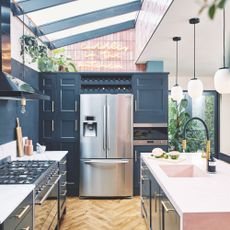 Kitchen with blue cabinets, wooden floors and a white worktop. A large fridge-freezer sits at the end of the kitchen