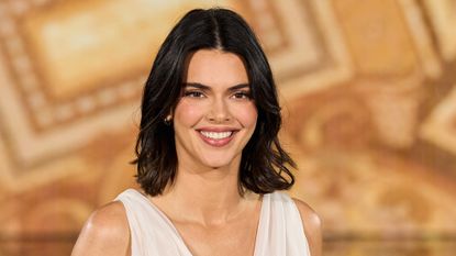 Kendall Jenner walks the runway wearing a white dress in front of a gold background