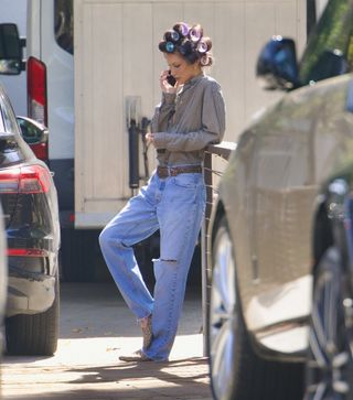 Bella Hadid lounges on set while wearing relaxed denim jeans with ripped knees, cheetah print loafers, and a baggy shirt