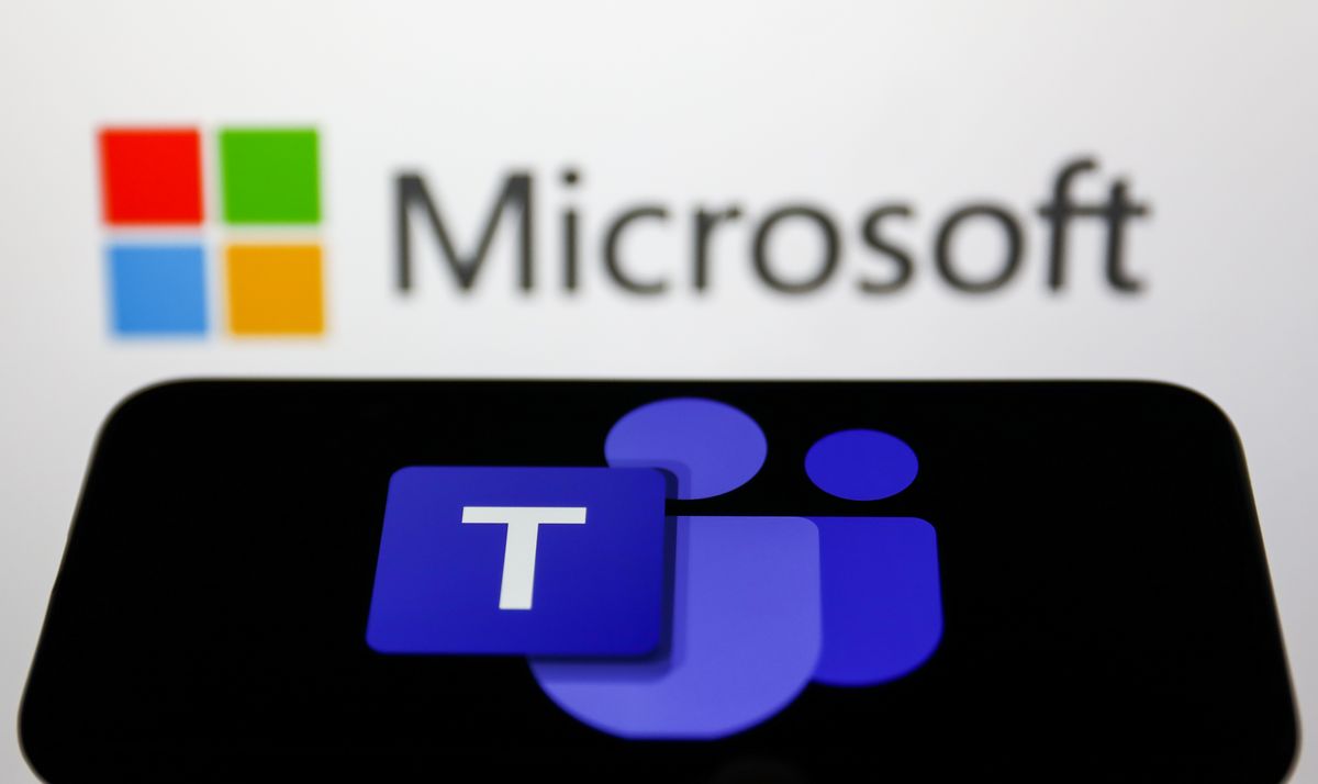 The Microsoft teams icon on a phone in the foreground, with the Microsoft logo out of focus against white in the background,