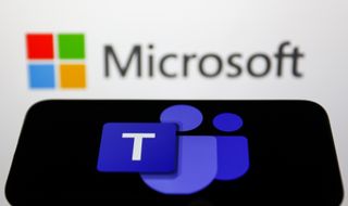 The Microsoft teams icon on a phone in the foreground, with the Microsoft logo out of focus against white in the background,