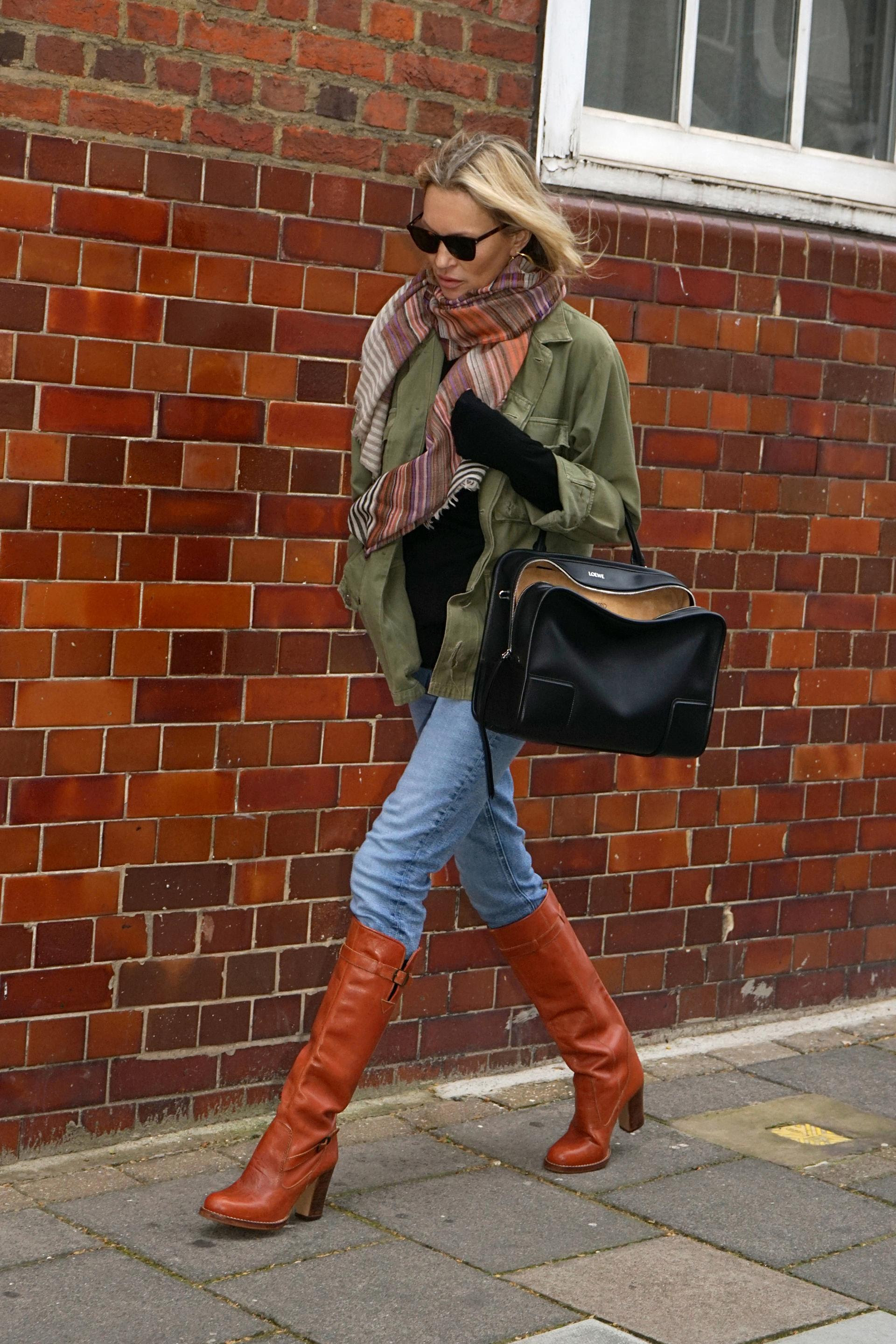 Kate Moss walks down the street wearing skinny jeans and knee-high boots with a khaki jacket and oversized scarf.