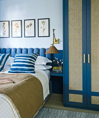 Pale blue painted bedroom, indigo scalloped headboard, wardrobe with rattan doors in matching indigo shade, white bed linen with white and indigo striped cushions, brown throw, matching artwork above the bed showing pressed flowers, small bedside shelf, wall light with woven wicker shade above.
