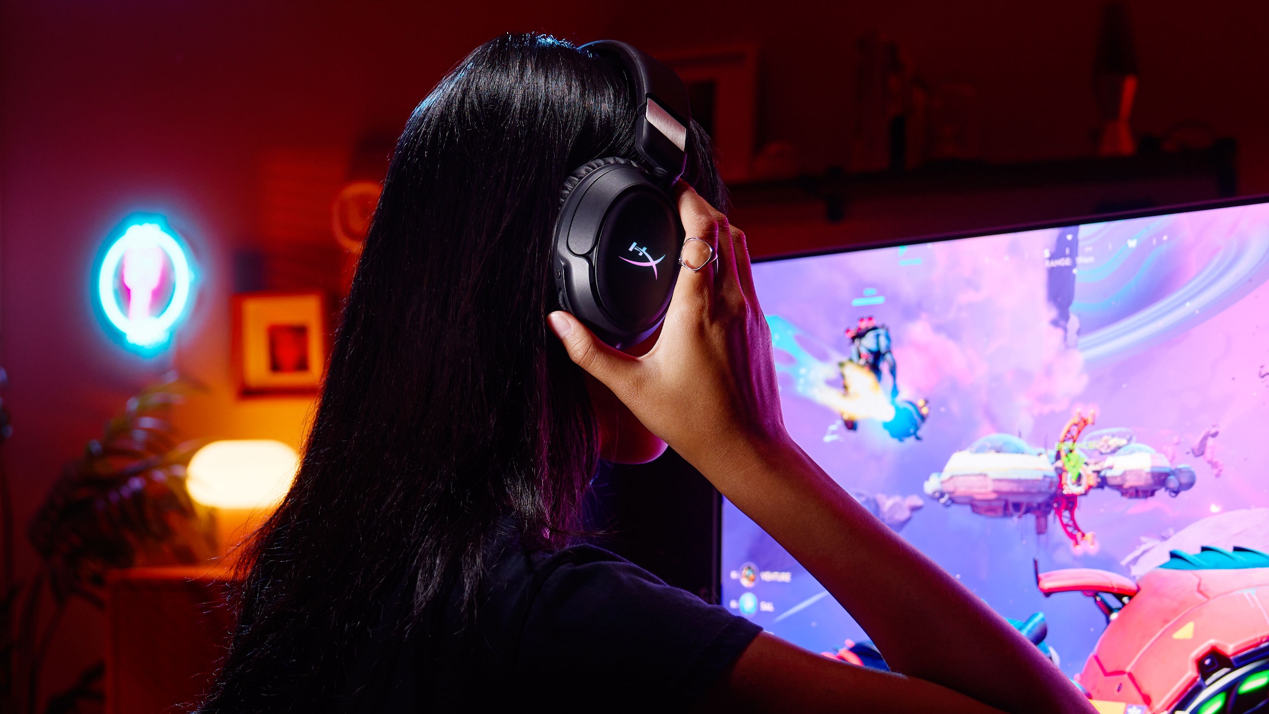 A person with long dark hair sits in a gaming room with a montior and adjusts a HyperX wireless gaming headset.