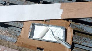Mini foam roller and tray with white paint resting on cardboard with a half-painted plywood shelf next to it