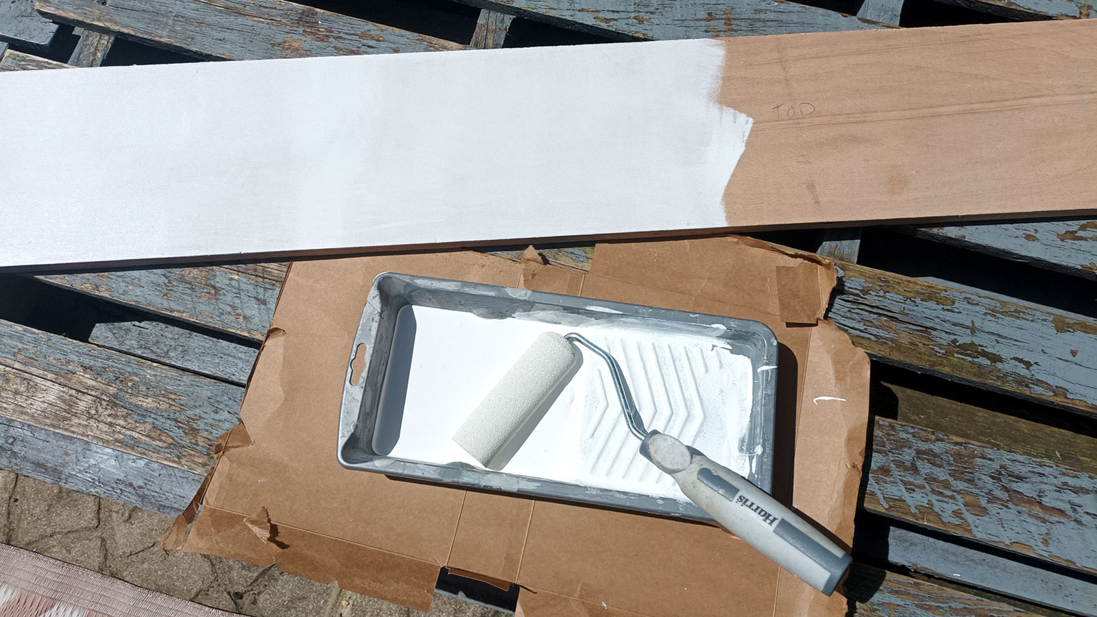 Mini foam roller and tray with white paint resting on cardboard with a half-painted plywood shelf next to it 