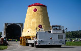 a cone-shaped rocket piece on a vehicle being moved towards an open storage container mounted on a ship