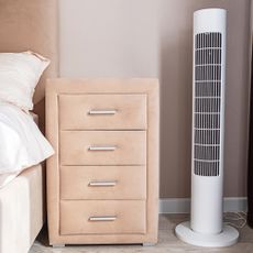White tower fan on the floor near a bedside table next to a bed with a padded headboard