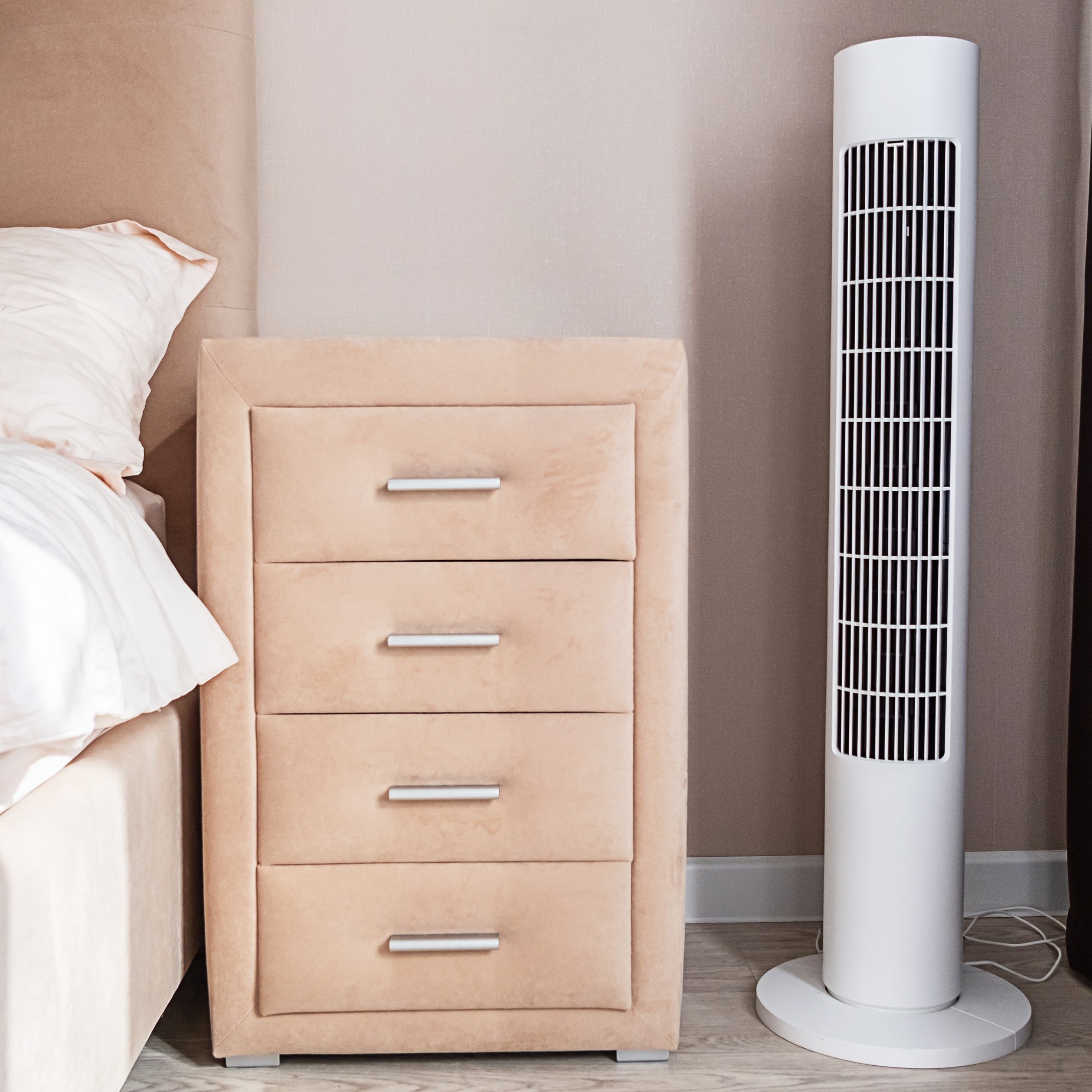 White tower fan on the floor near a bedside table next to a bed with a padded headboard