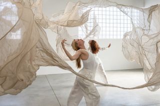 Two young female dancers dressed in white tops and trousers dance around a textile, large-scale sculpture floating in a gallery space filled with natural light.