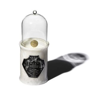 The best candle for homes, this Officine Universelle Buly is on a white, marble base with a glass dome