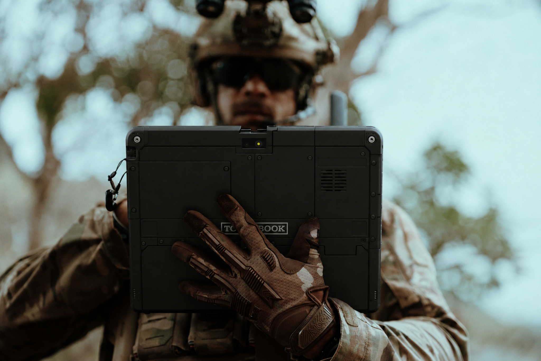 A soldier in tactical gear and gloves is holding the back of a rugged Panasonic TOUGHBOOK 34 tablet outdoors.