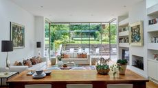 A bright and sunny kitchen diner with large windows showing a well-kept garden behind. In the foreground is a large wooden table, and seen behind is a comfy seating area with white sofas, a patterned rug, and large bookshelves. Colorful abstract art is seen framed on the walls.