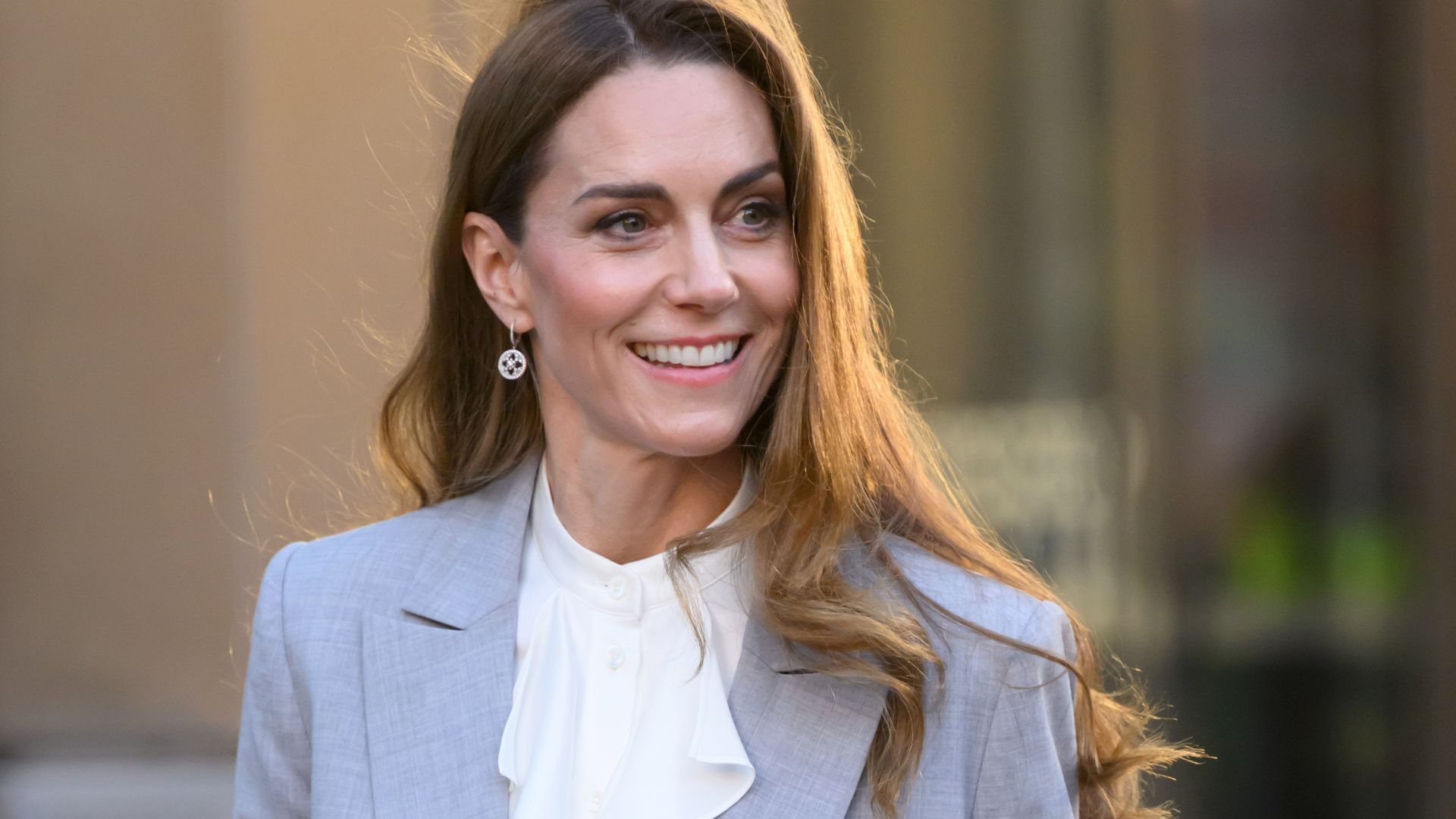 Kate Middleton steps outside wearing a light grey suit with black pointed-toe heels and a cream ruffle blouse.