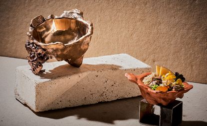 A bronze circular dish with rugged edges sat on a brick. Below, a smaller dish filled with oatmeal and other pulses. 