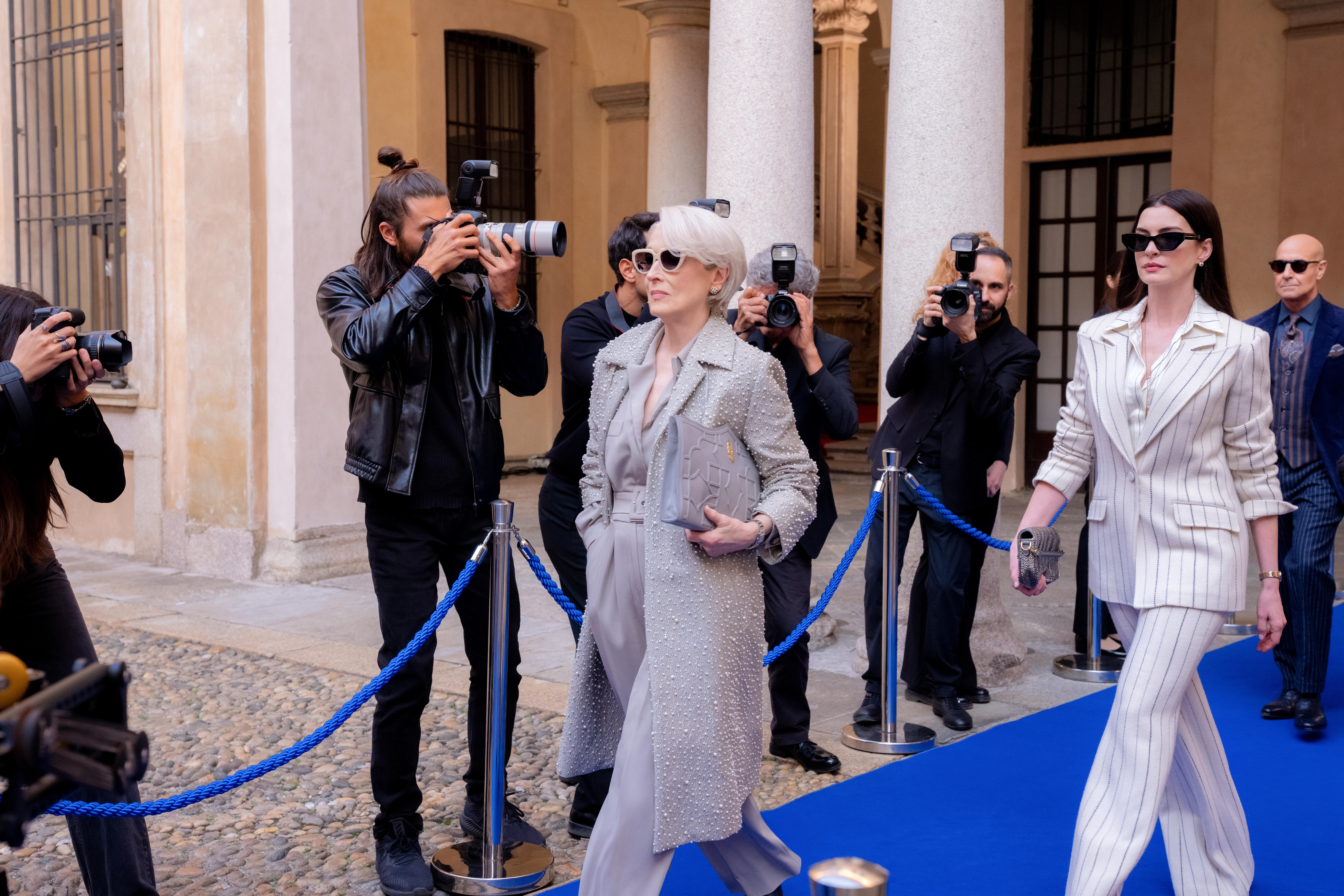 Meryl Streep as Miranda Priestly wearing a grey jacket Anne Hathaway as Andy Sachs wearing a striped suit walking down a carpet as photographers take photos in the devil wears prada 2