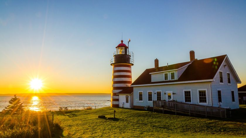 a sunrise on the left over water and a quaint lighthouse in the foreground on the right.