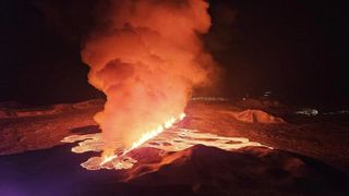 A river of lava erupting at night time with large plumes of ash