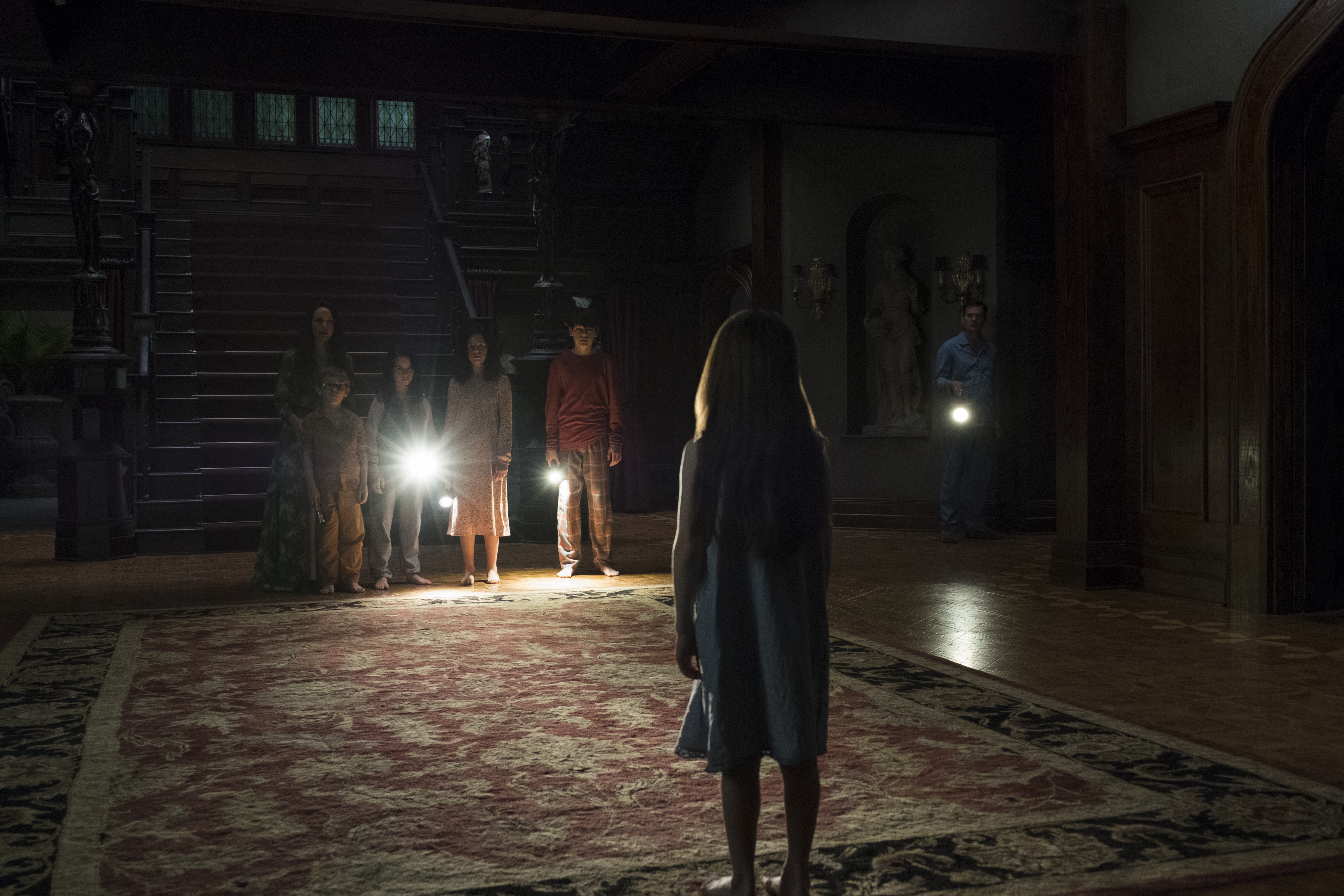 The Haunting of Hill House still featuring a little girl standing in the dark of a parlor and her family shining flashlights on her