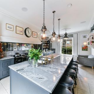 a grey and white large kitchen that has grey cupboards with marble tops, black bar stools, three chrome globe pendant lights hanging over the kitchen island and gold tap fixings on the double sink
