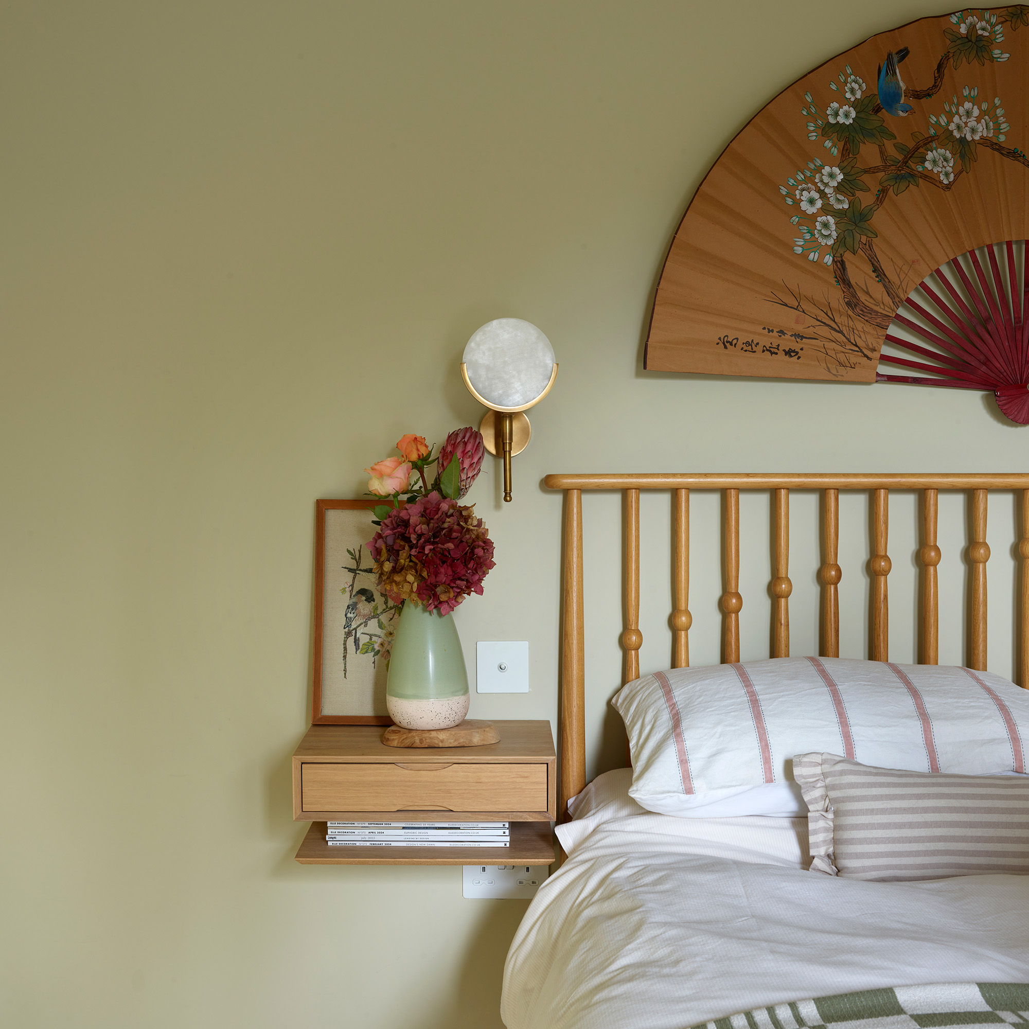 bedroom with green wall floating bedside table wall light and fan above a double bed with wooden frame and white bedlinen and green throw